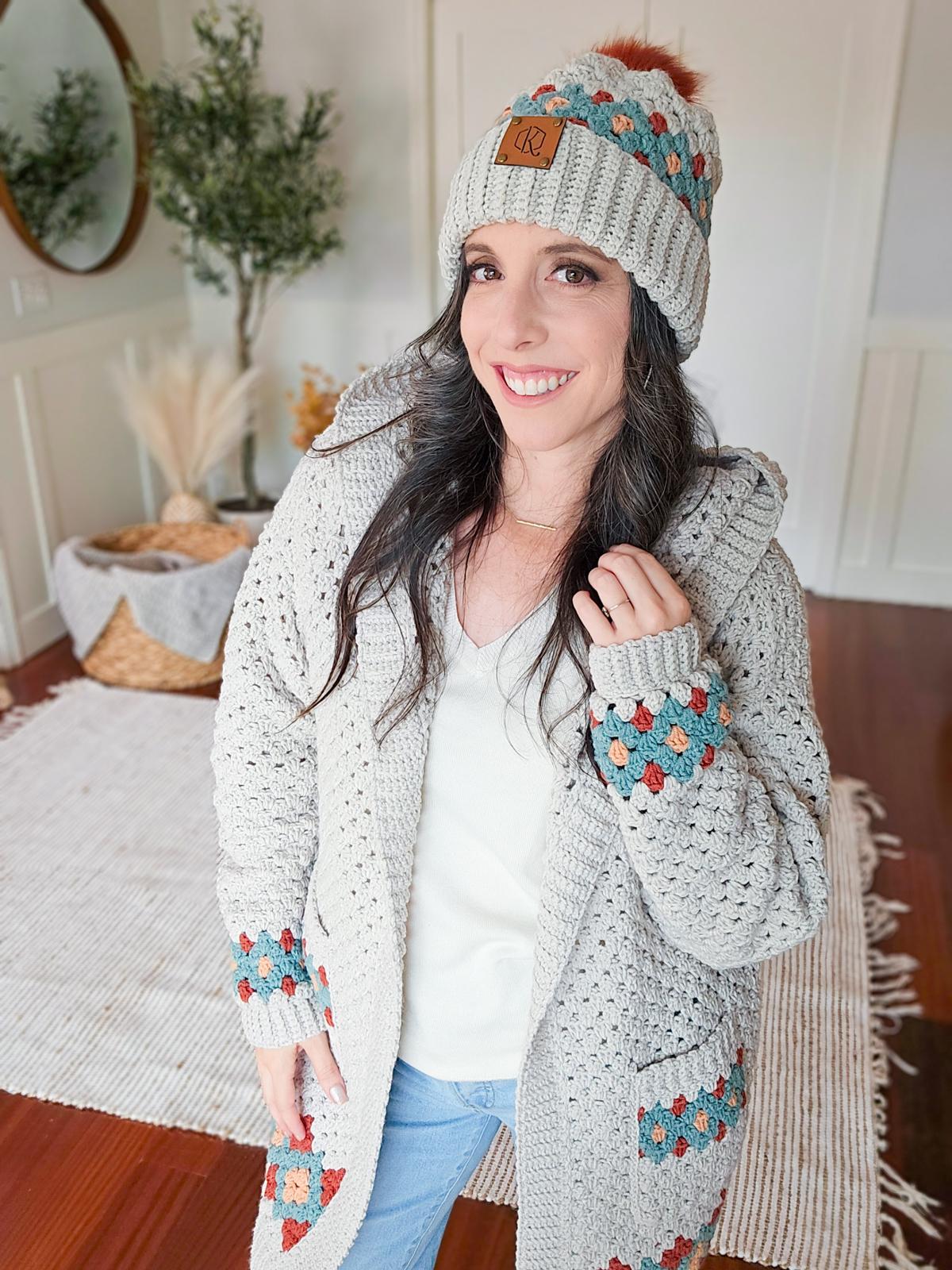 A woman in a crochet hat and cardigan stands indoors, smiling warmly. Her clothing, a handmade gift, showcases a gray and multicolored pattern. A rug and basket lend a cozy charm to the background.