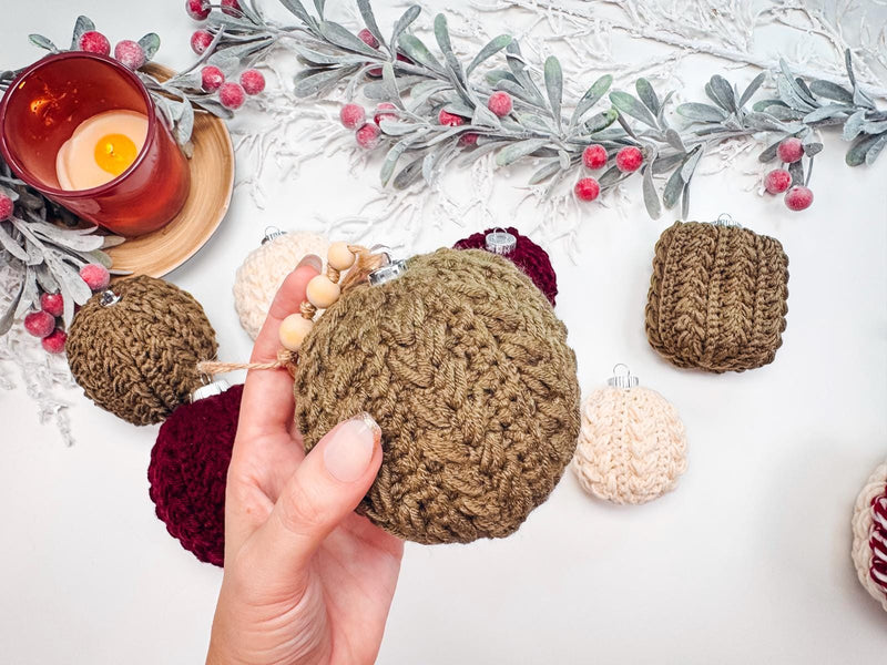 A hand holds a crochet Christmas ornament surrounded by other ornaments and holiday decor on a table, capturing the charm of a handmade gift.