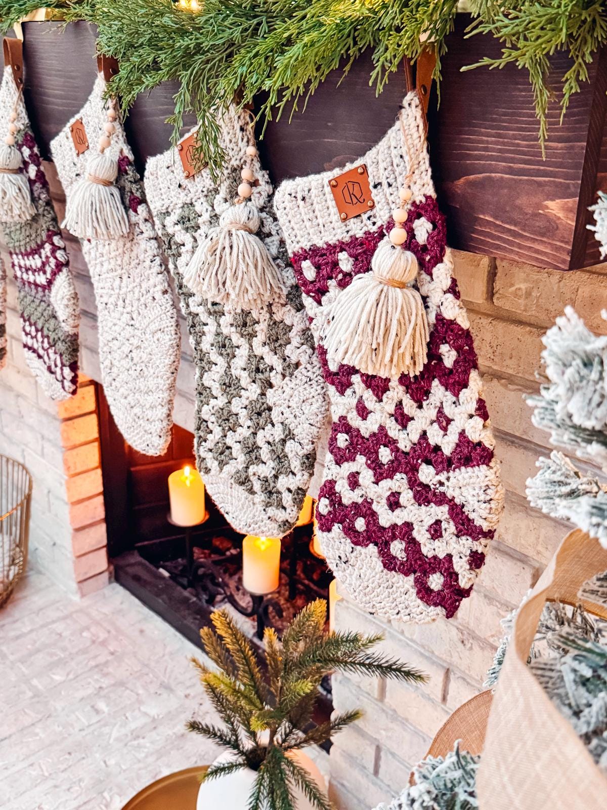 Crocheted Christmas stockings, resembling vintage clothes, hang on a decorated mantel with candles and a small tree nearby. These handmade gifts add a touch of timeless charm to the festive setting.
