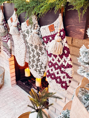 Crocheted Christmas stockings, resembling vintage clothes, hang on a decorated mantel with candles and a small tree nearby. These handmade gifts add a touch of timeless charm to the festive setting.