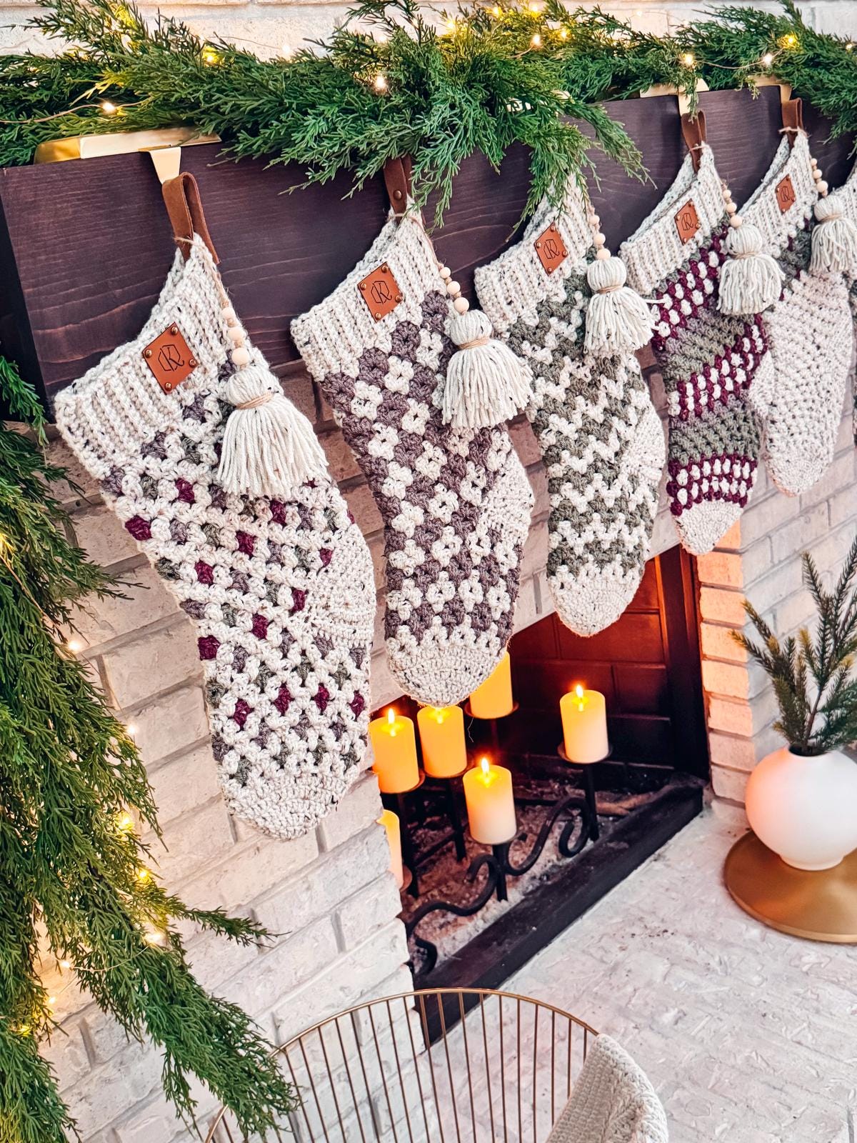 Crocheted stockings with tassels hang from a mantel decorated with greenery and lights above a lit fireplace, evoking the charm of handmade gifts.