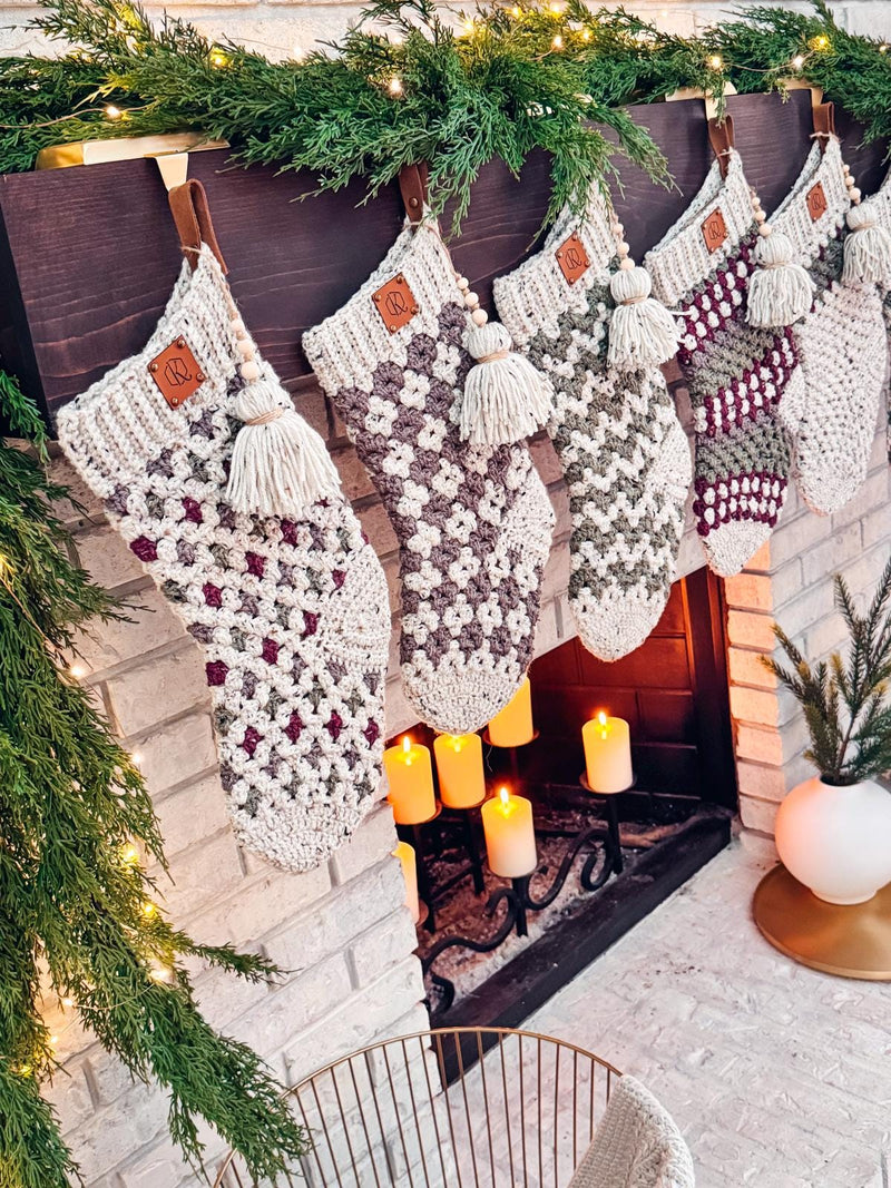 Crocheted stockings with tassels hang from a mantel decorated with greenery and lights above a lit fireplace, evoking the charm of handmade gifts.