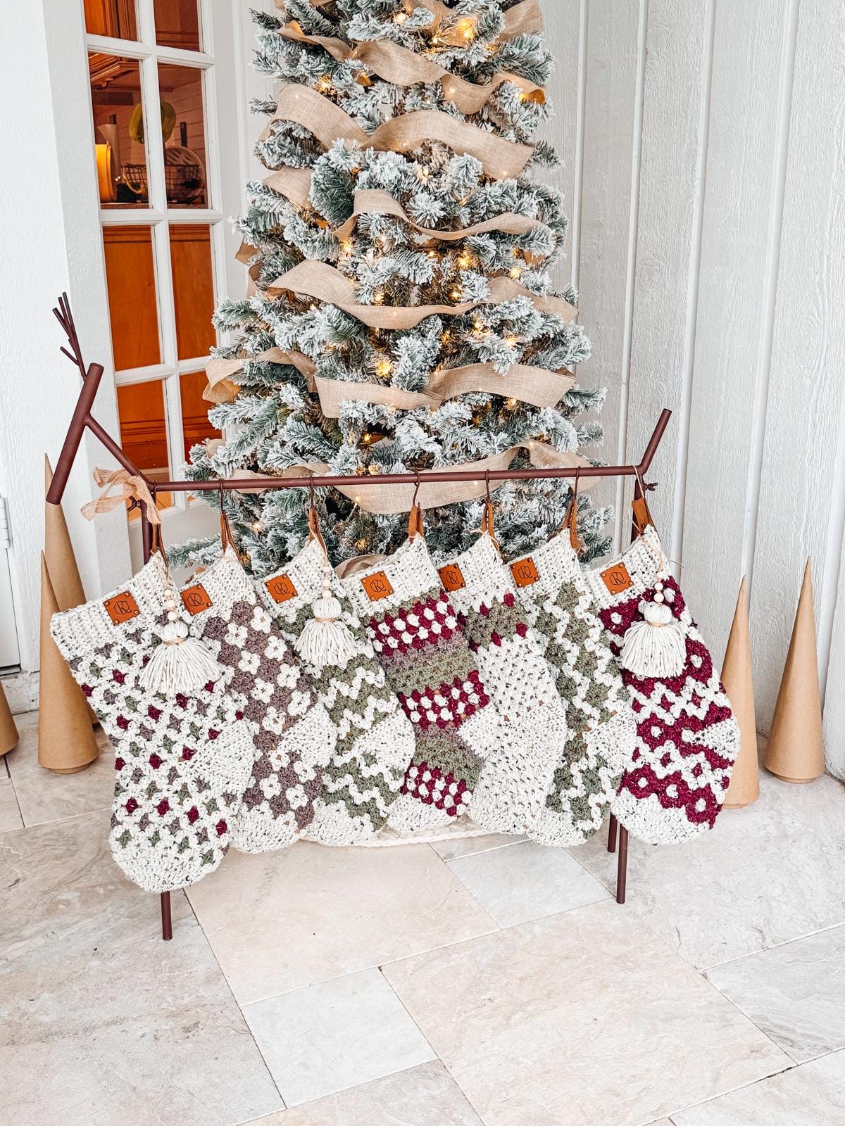 Seven handcrafted crochet stockings with various patterns hang in front of a decorated, flocked Christmas tree indoors, offering the warmth of a handmade gift.