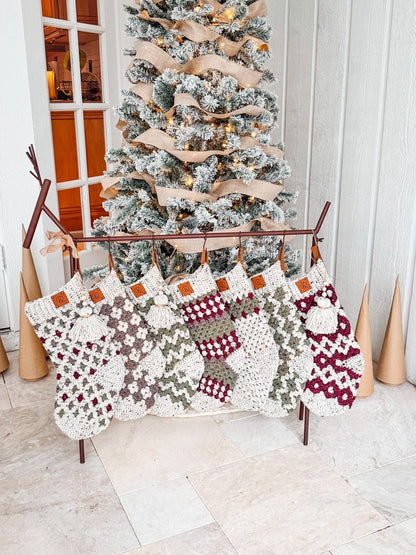 Seven handcrafted crochet stockings with various patterns hang in front of a decorated, flocked Christmas tree indoors, offering the warmth of a handmade gift.