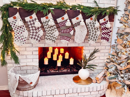A brick fireplace with lit candles sits beneath a mantle draped in garland and seven crochet stockings. Nearby, a Christmas tree stands adorned, next to a basket holding a cozy blanket and handmade gifts.
