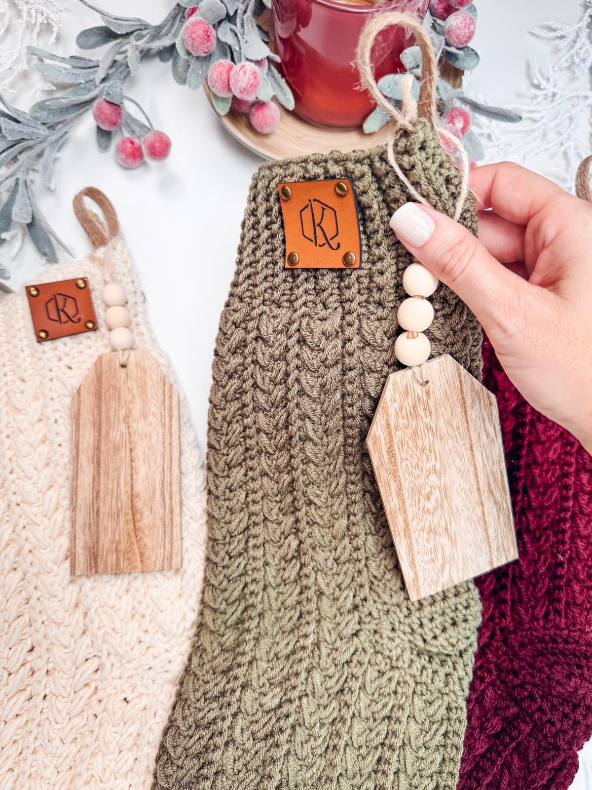 Hand holding a green crochet stocking with a wooden tag, next to a cream and burgundy stocking. This handmade gift complements holiday decor with frosted leaves and berries in the background, adding a touch of timeless charm.