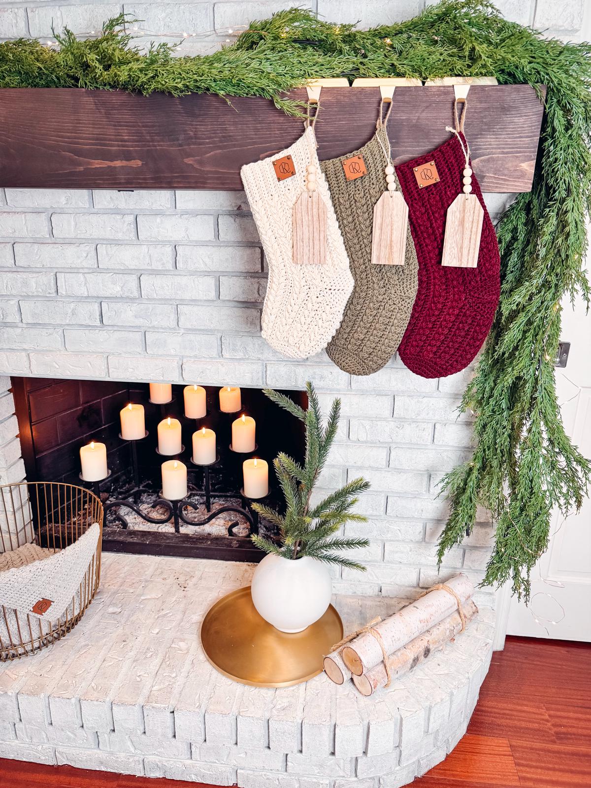 A brick fireplace is adorned with green foliage and three crochet stockings. Candle lights illuminate the space, while a plant, birch logs, and a handmade gift add charm to the hearth.
