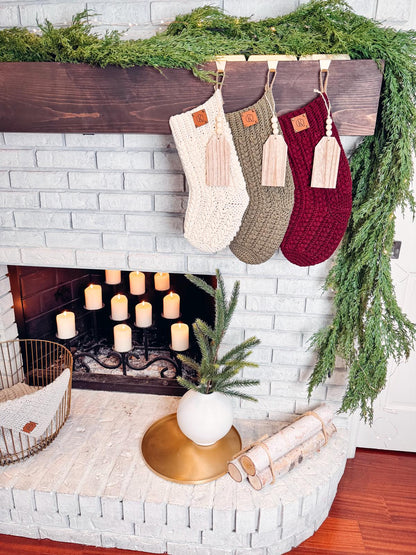 A brick fireplace is adorned with green foliage and three crochet stockings. Candle lights illuminate the space, while a plant, birch logs, and a handmade gift add charm to the hearth.