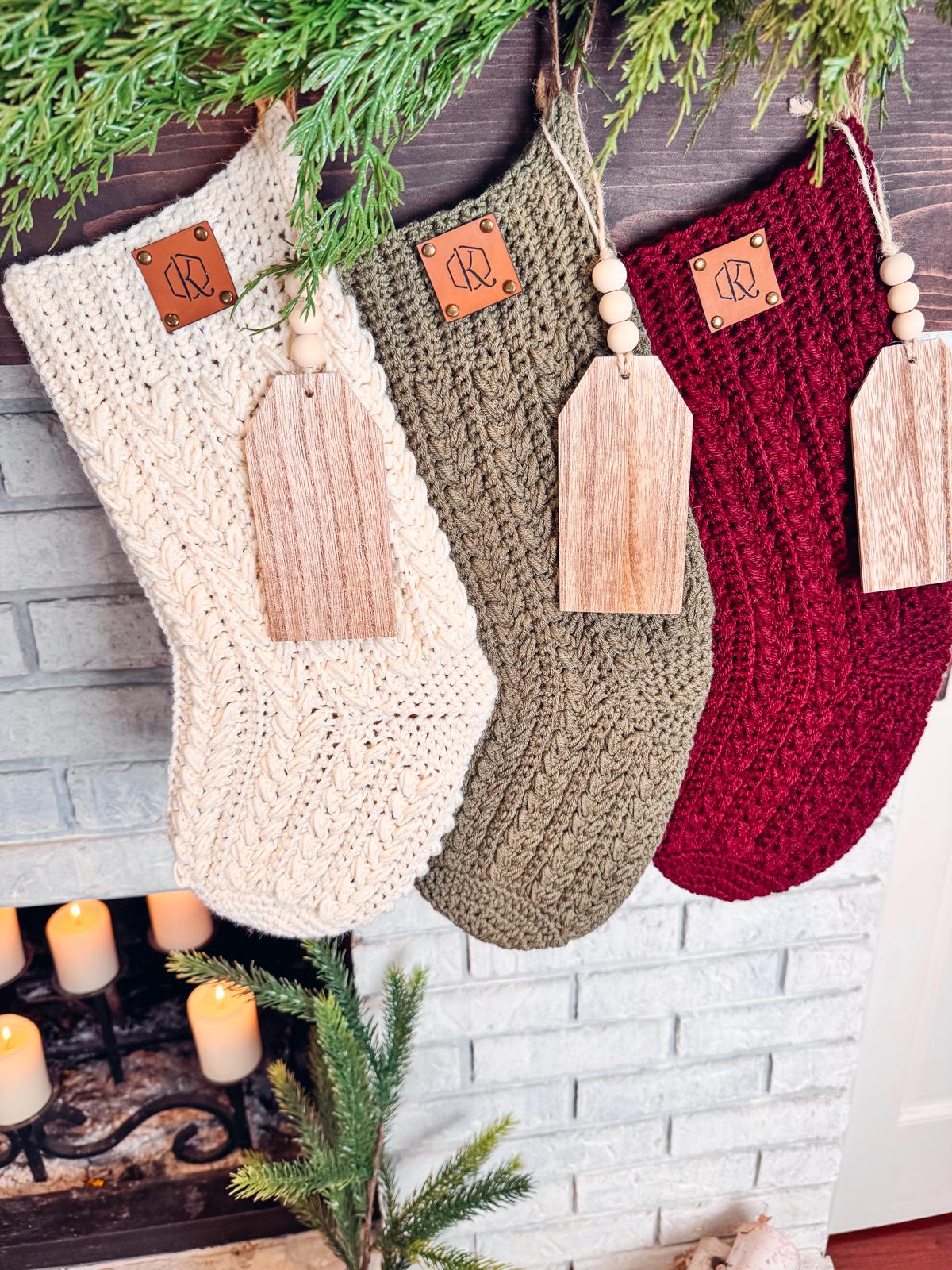 Three crocheted Christmas stockings in cream, green, and red hang by a fireplace. Each, resembling handmade gifts, features a wooden tag and decorative branch above.