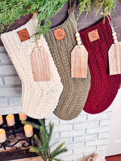 Three crocheted Christmas stockings in cream, green, and red hang by a fireplace. Each, resembling handmade gifts, features a wooden tag and decorative branch above.