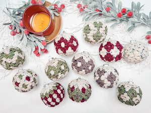 Crocheted holiday ornaments in red, green, and white patterns are arranged on a table with faux greenery, a cup of tea, and the perfect touch for a handmade gift.
