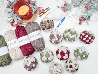 Various crochet Christmas ornaments and yarn balls are arranged on a table with crafting tools, a lit candle, and decorative greenery, making it the perfect setting for creating handmade gifts.