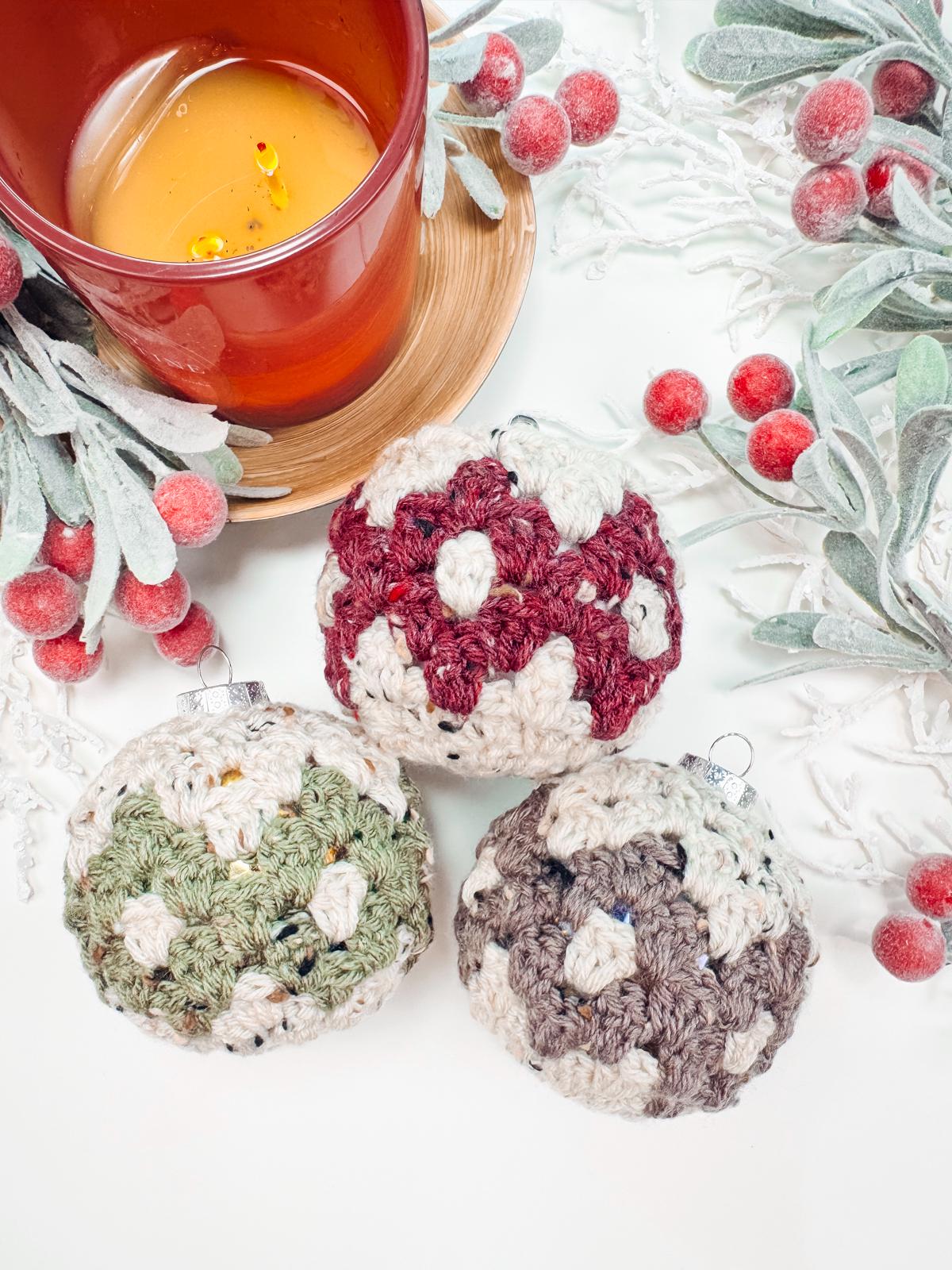 A red candle sits on a wooden coaster next to three crocheted baubles in red, green, and gray, surrounded by frosted leaves and red berries—an ensemble that makes for a charming handmade gift.