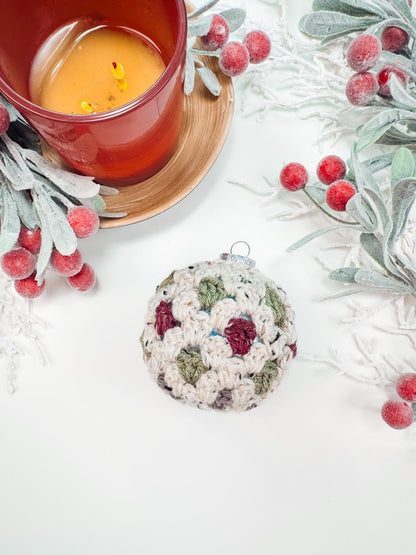 Crocheted ornament with multicolored patterns next to a lit orange candle in a wooden holder, surrounded by frosted greenery and red berries on a white surface—a perfect handmade gift for those who appreciate artisanal charm.