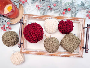 A wooden tray holds handmade crochet ornaments in red, cream, and green. A candle and frosted leaves with red berries are beside it on a white surface, creating the perfect scene for a thoughtful handmade gift.