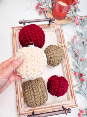 A hand holds a crochet ornament above a tray filled with red, green, and cream knitted ornaments, perfect as a handmade gift. Nearby, a candle flickers softly amidst decorative foliage.