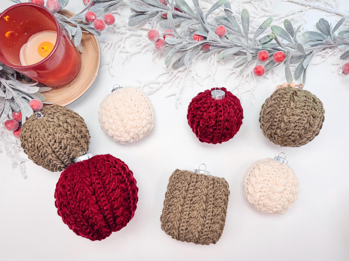 A decorative arrangement of handmade rocheted ornaments in red, brown, and cream alongside a red candle and frosted leaves with berries on a white surface. Perfect as a thoughtful gift.
