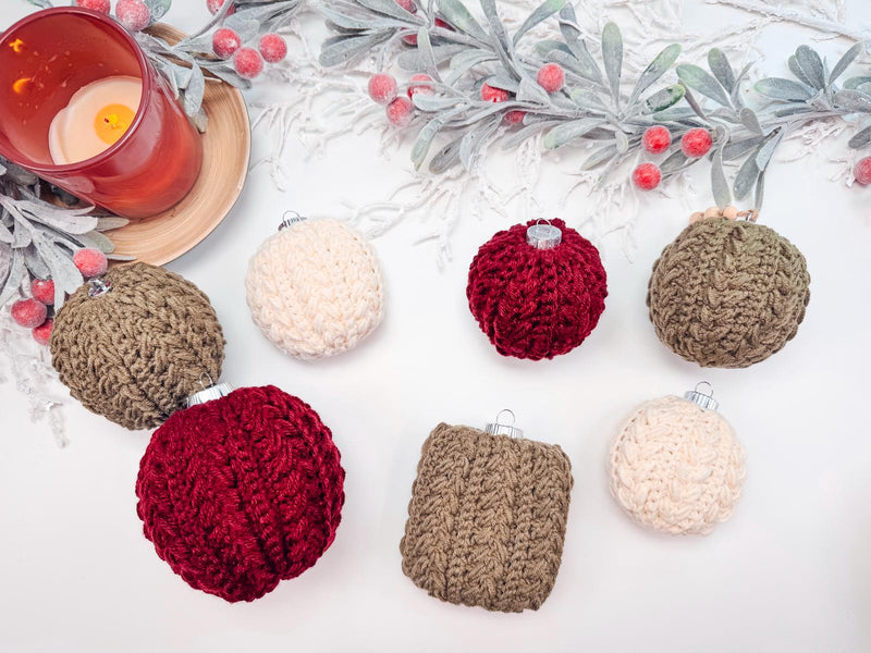 A decorative arrangement of handmade rocheted ornaments in red, brown, and cream alongside a red candle and frosted leaves with berries on a white surface. Perfect as a thoughtful gift.