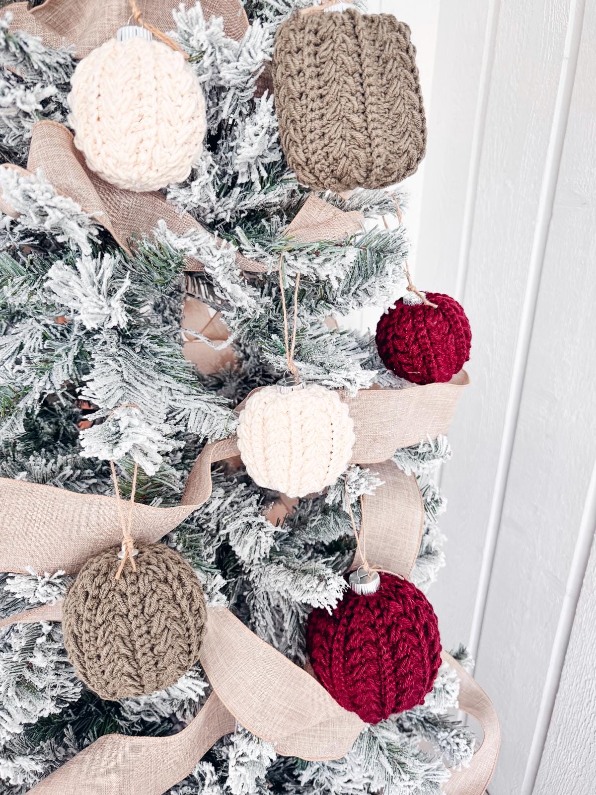 Christmas tree with crocheted ornaments in cream, brown, and red, adorned with a beige ribbon around snowy branches, adds the charm of a handmade gift.