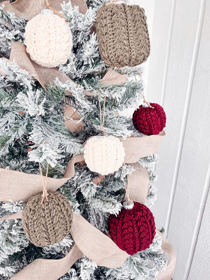 Christmas tree with crocheted ornaments in cream, brown, and red, adorned with a beige ribbon around snowy branches, adds the charm of a handmade gift.