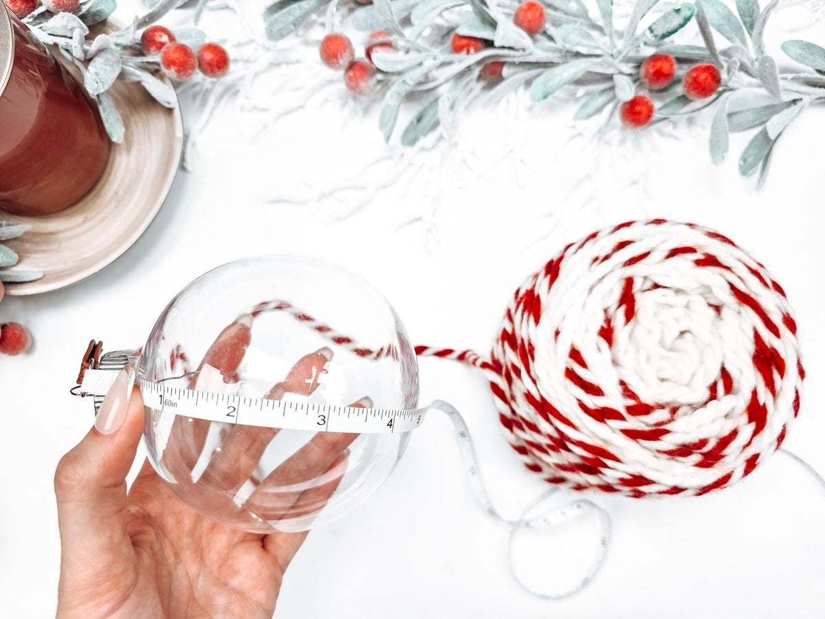 A hand holds a clear ornament next to handmade gift-wrapped balls of red and white yarn and a tape measure, with decorative foliage and berries in the background.