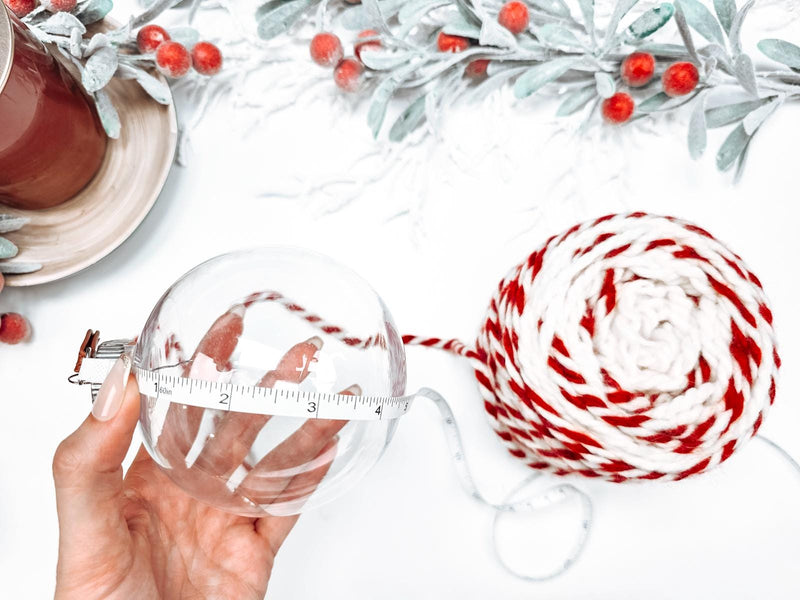 A hand holds a clear ornament next to handmade gift-wrapped balls of red and white yarn and a tape measure, with decorative foliage and berries in the background.