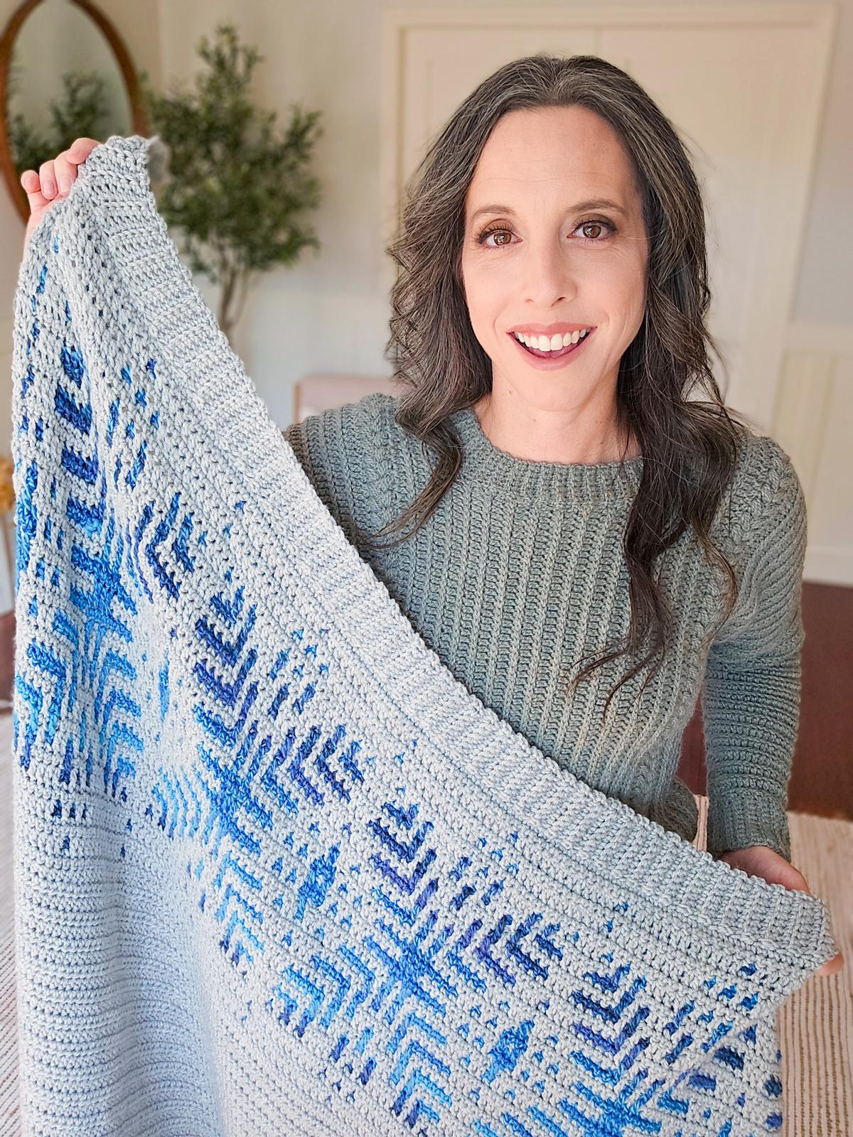 A person smiling, holding up a crocheted blanket with blue geometric snowflake patterns, standing indoors—an ideal handmade gift to complement your collection of unique jewellery.