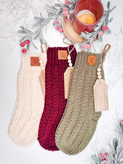 Three crochet Christmas stockings in cream, red, and green with wooden tags are adorned with faux frosted branches and berries next to a lit candle in a red holder, making a charming handmade gift for those who appreciate artistry and seasonal cheer.
