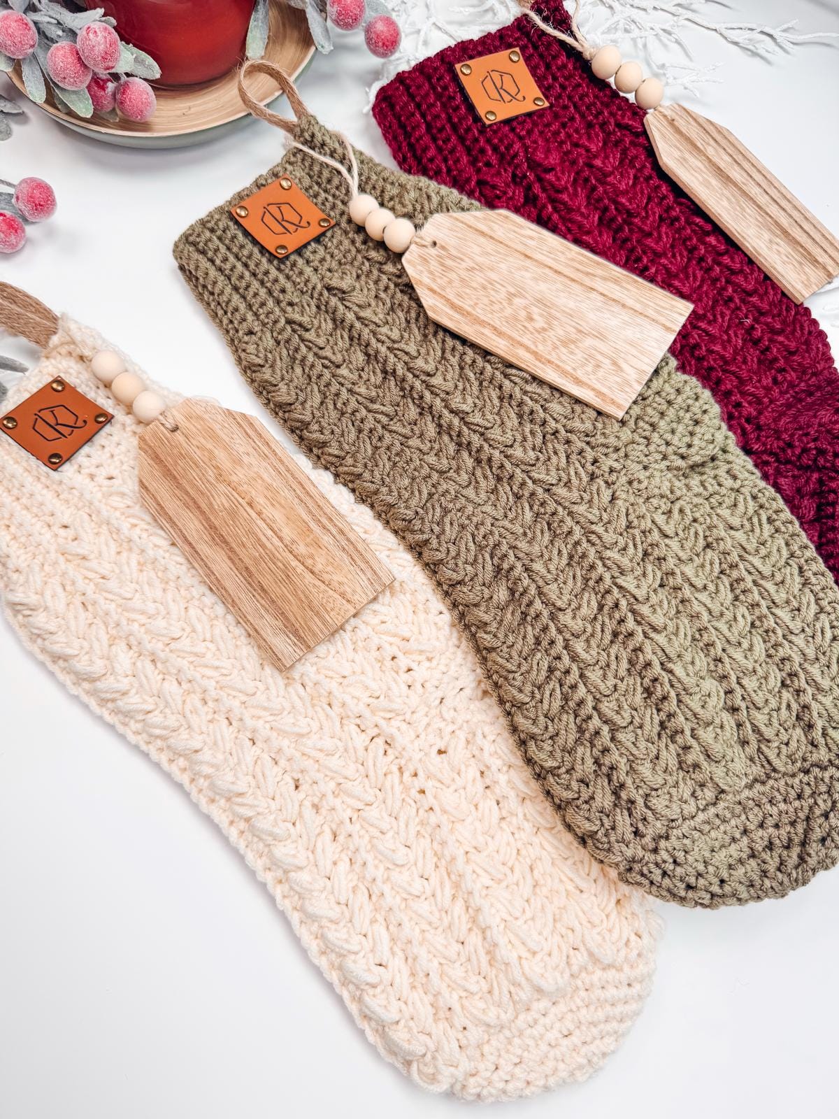 Three crochet stockings in cream, olive green, and burgundy are placed in a row, each adorned with a wooden name tag and leather patch. Perfect as a handmade gift, these stockings add a touch of charm and warmth to any holiday décor.