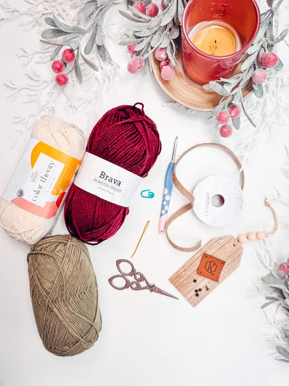 Yarn balls, crochet hook, crochet hook, scissors, ribbon, wooden tags, stitch marker, candle, and decorative branches on a white surface create the perfect setting for crafting a handmade gift.