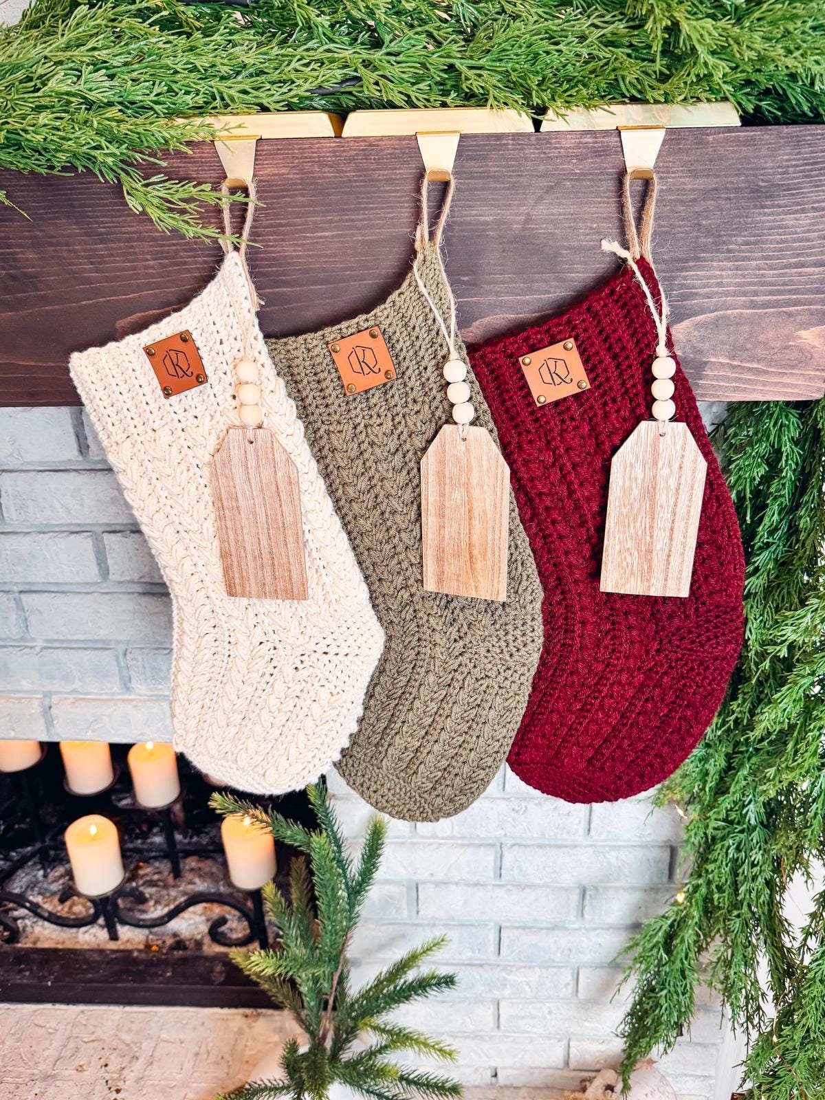 Three crocheted Christmas stockings in cream, green, and red hang on a wooden mantle decorated with greenery. Above an unlit white brick fireplace with candles, this charming scene evokes the warmth of a handmade gift.