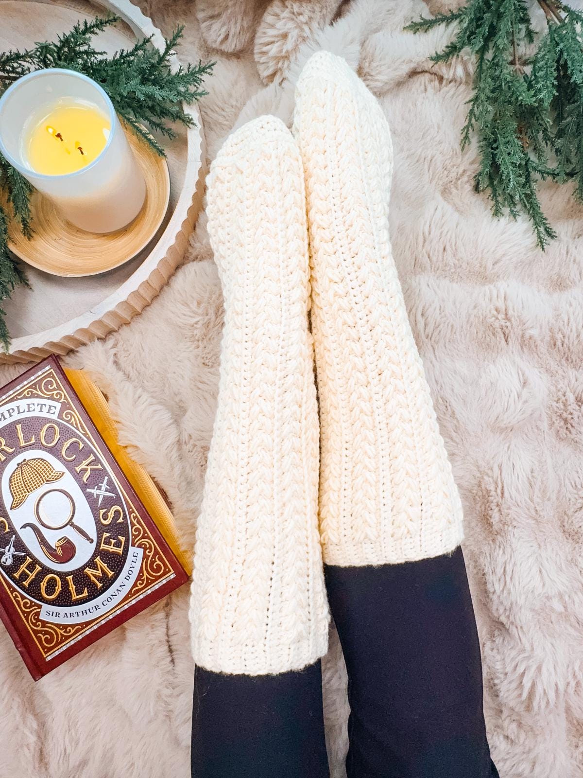 A cozy scene unfolds with a person wearing cream crocheted slipper socks, lounging on a beige furry blanket beside the soft glow of a candle. Nearby rests a book titled "Sherlock Holmes", adding a touch of elegance to this tranquil moment.