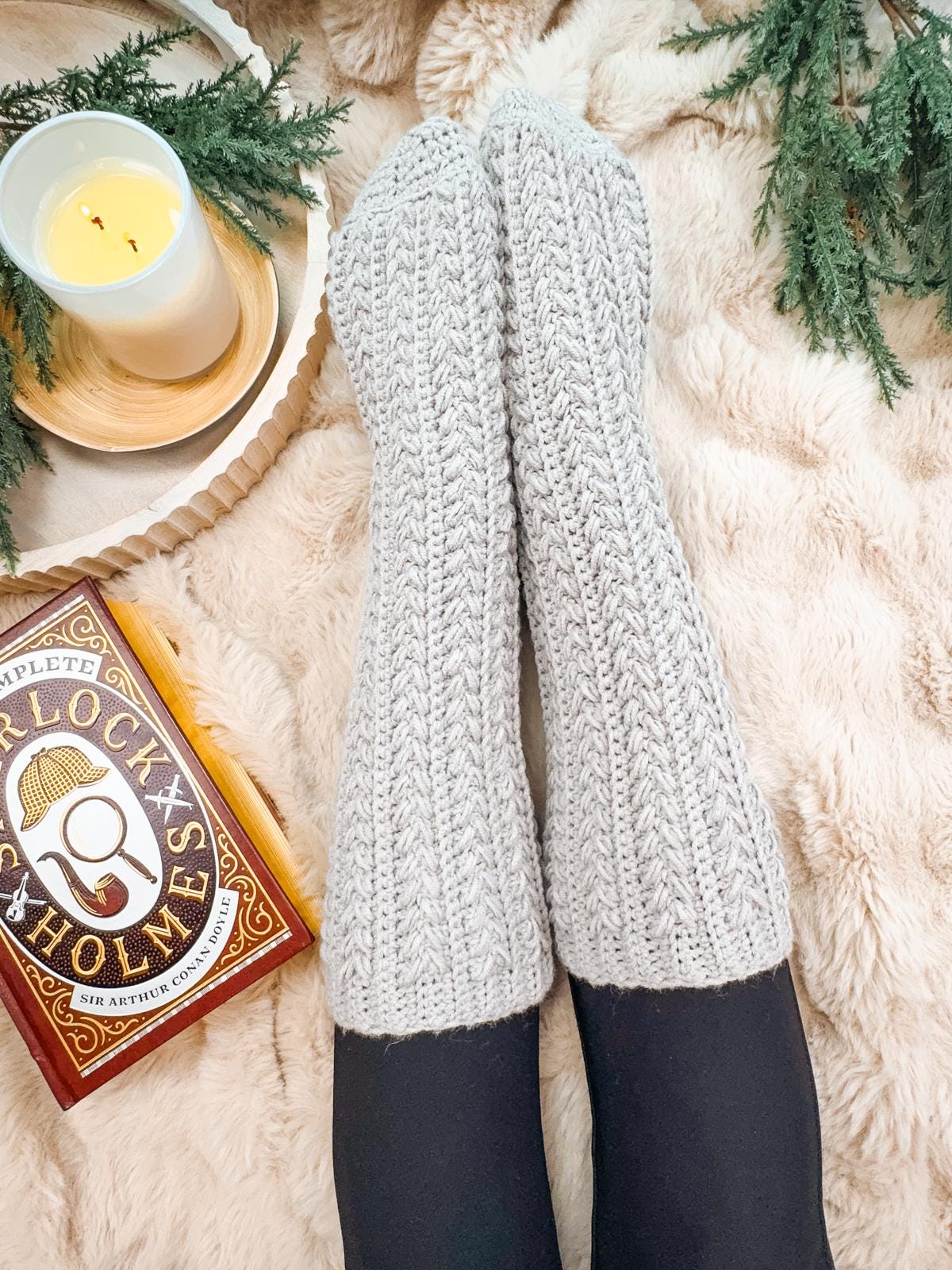 Feet in gray crocheted slipper socks rest on a fluffy blanket, with a lit candle and a Sherlock Holmes book nearby, surrounded by greenery and hints of vintage clothes for style inspiration.