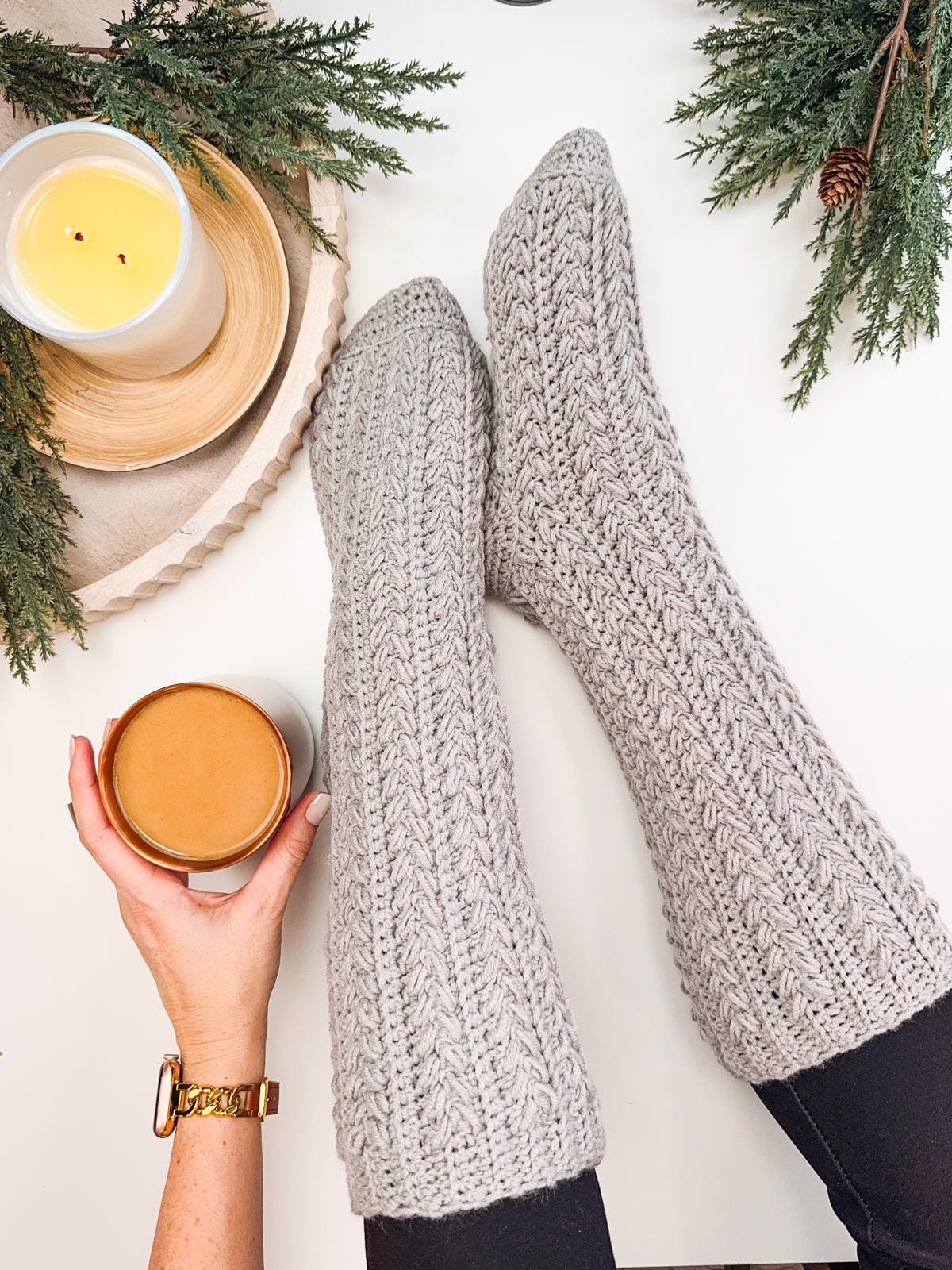 A person wearing gray crochet socks, holding a cup of coffee. Nearby, a lit candle, pine branches, and a handmade gift rest on the white surface, adding warmth and charm to the cozy scene.