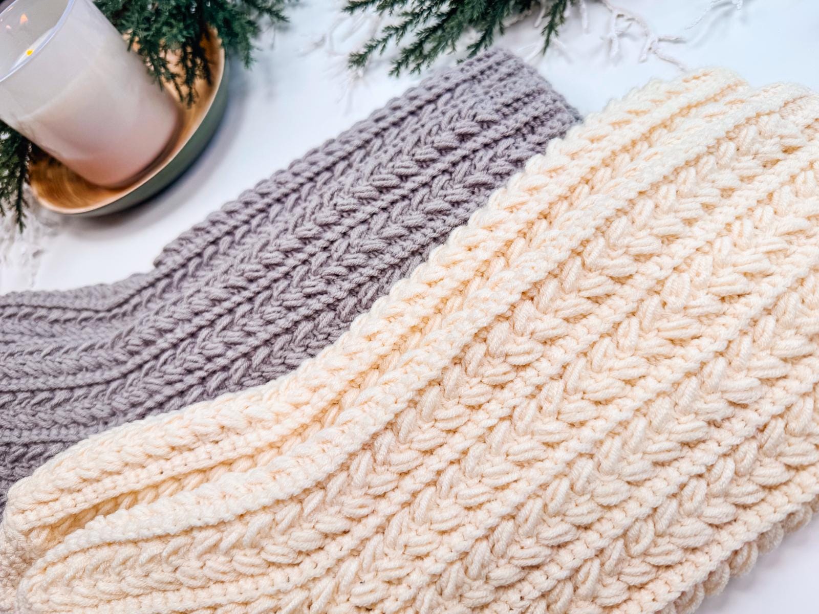 Crocheted beige and gray scarves rest on a table beside a lit candle and green pine branches, presenting the perfect handmade gift for loved ones.