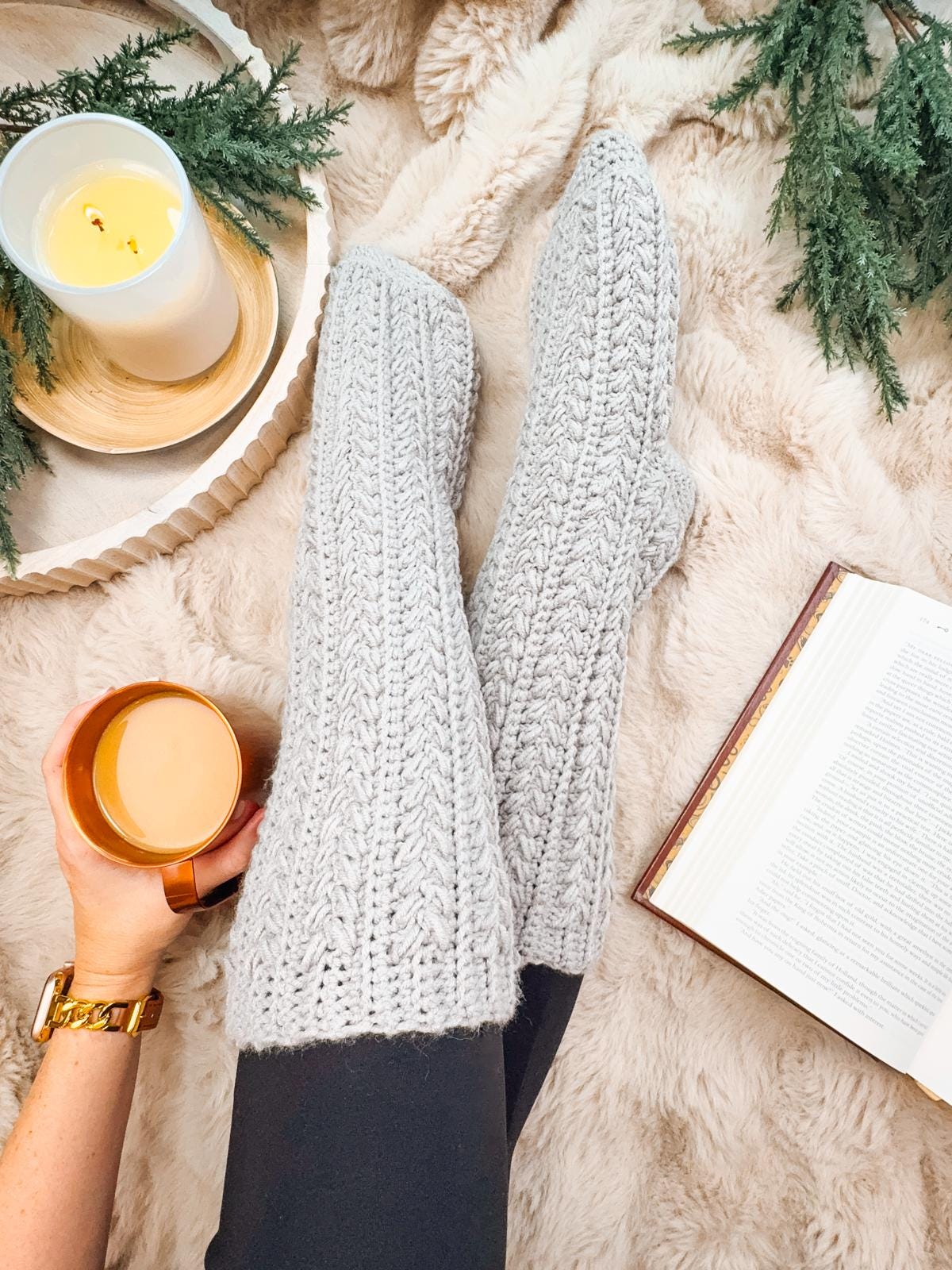 A cozy scene unfolds with a person wearing gray crochet socks, holding a coffee mug near an open book on a fluffy rug. A lit candle casts a warm glow, complemented by green branches and adorned with unique jewelry that adds a touch of elegance.
