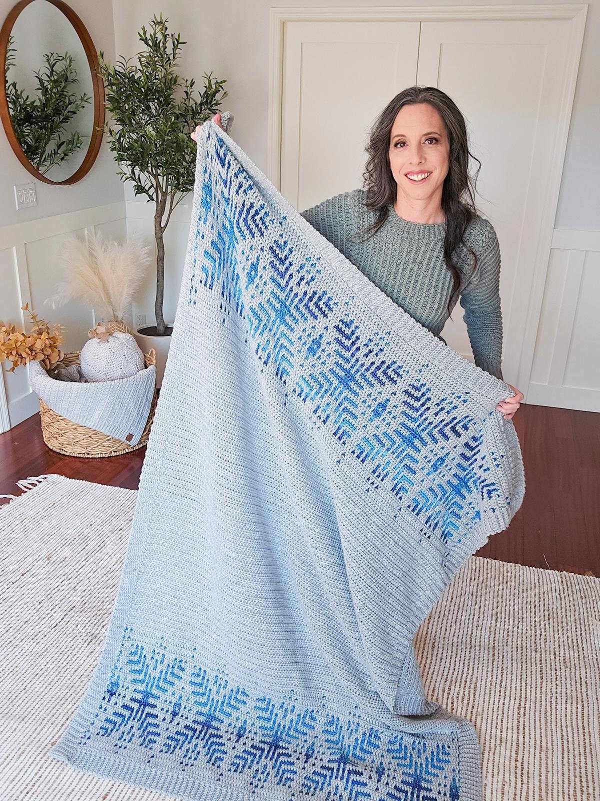 A person smiles while holding a large crochet gray blanket with blue snowflake patterns in a cozy living room, embracing the warmth of a handmade gift.