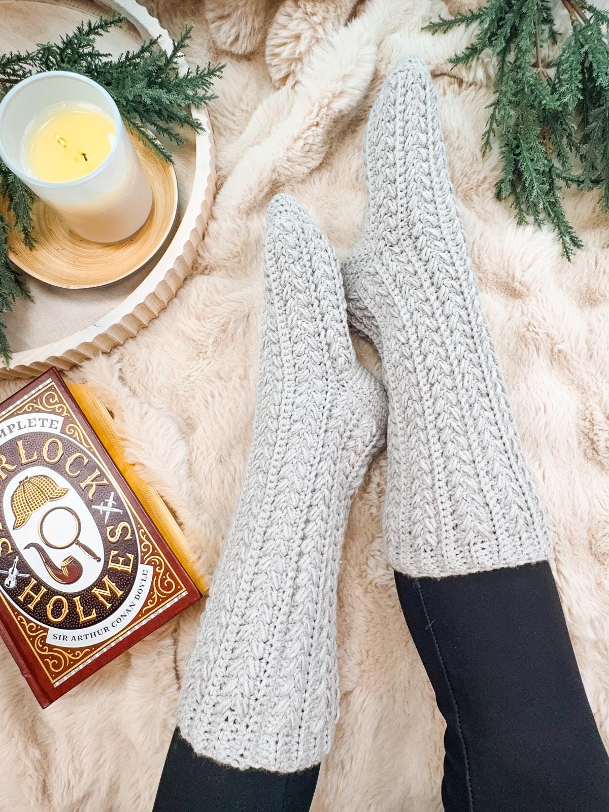 Feet in gray crocheted slipper socks rest on a fluffy blanket, with a lit candle and a Sherlock Holmes book nearby, surrounded by greenery and hints of vintage clothes for style inspiration.