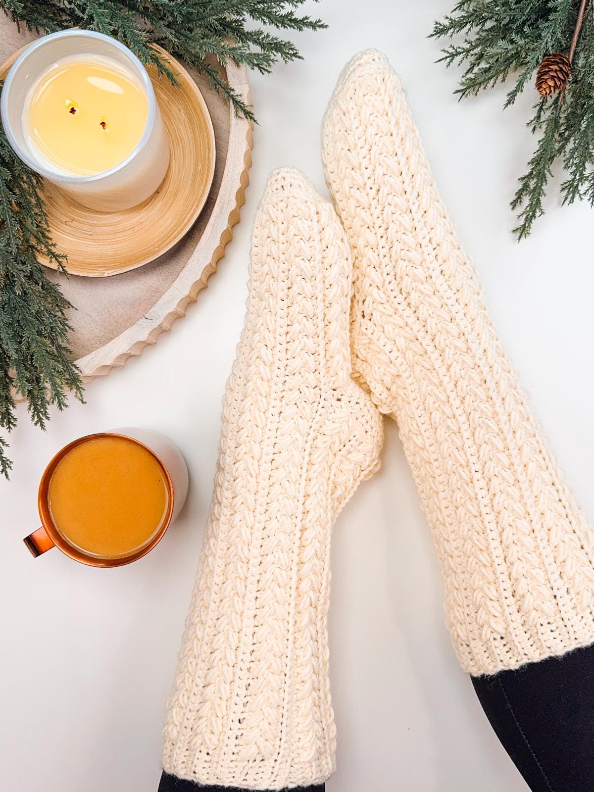 A person wearing cream crochet socks enjoys a cozy moment surrounded by green foliage on a white surface, with a lit candle and a cup of coffee. The scene exudes charm, perfect for those who appreciate handmade gifts.