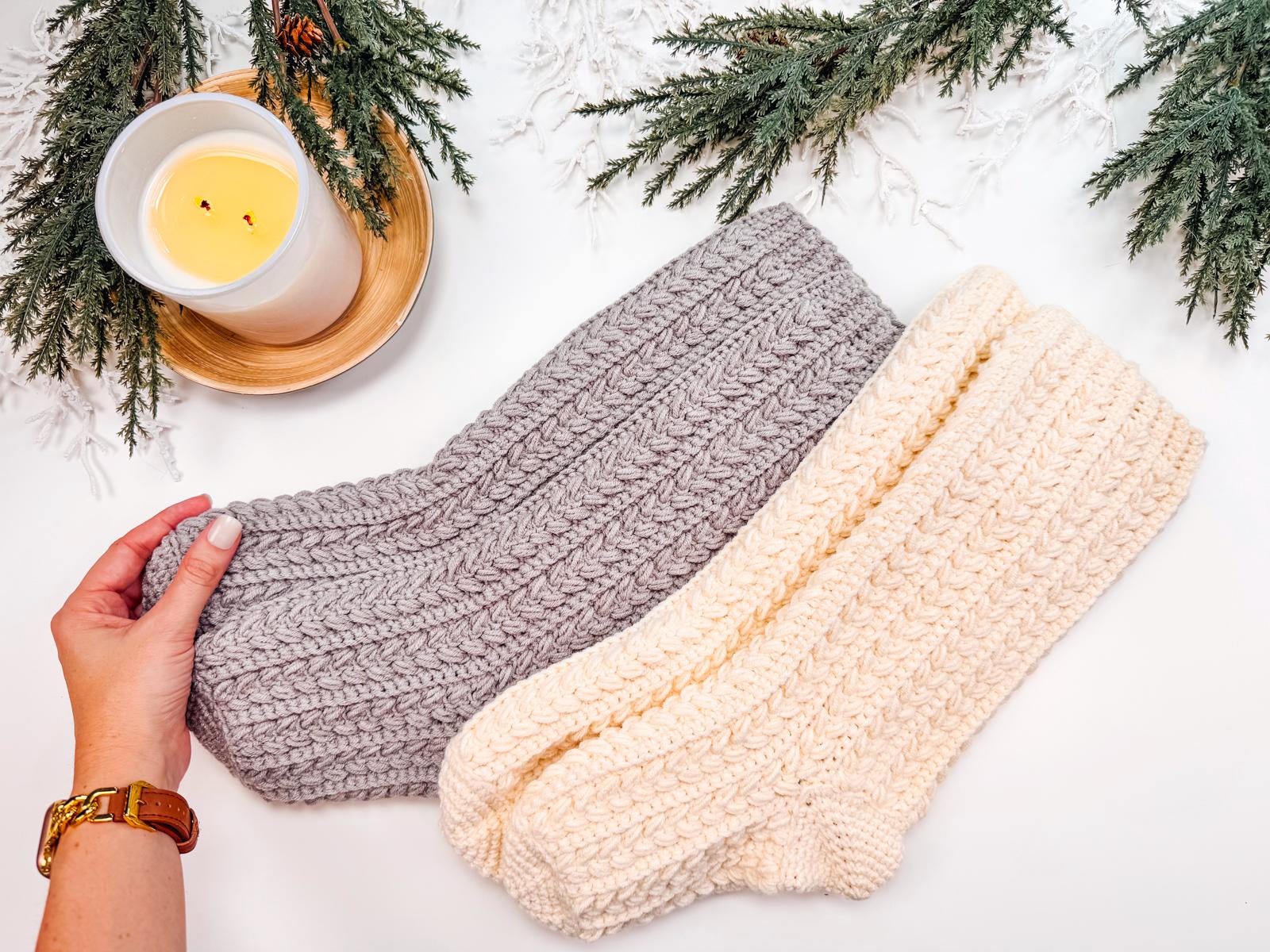 A hand gently touches gray and cream crocheted slipper socks on a table adorned with a lit candle and green pine branches, creating the perfect setting for a thoughtful handmade gift.