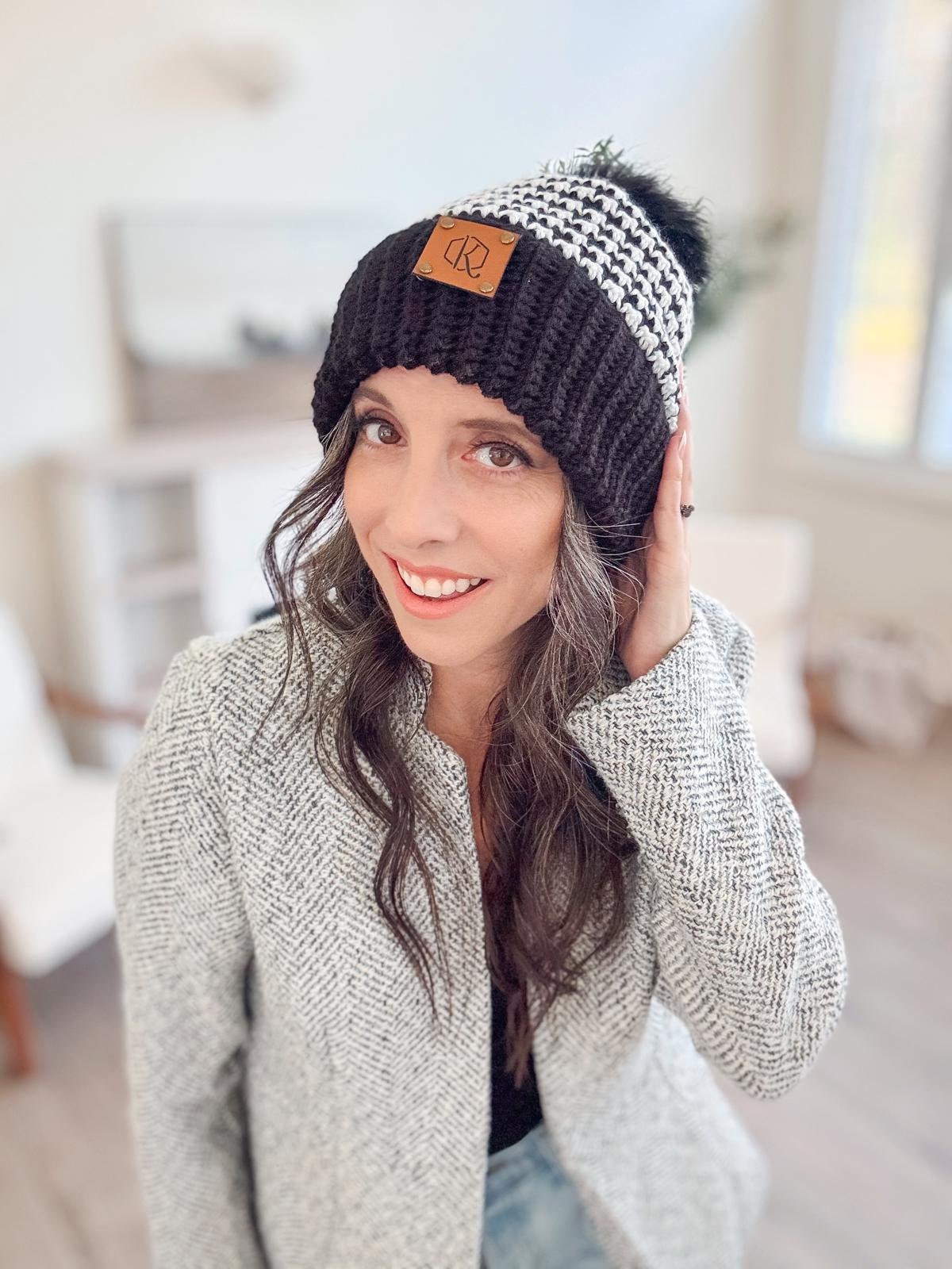 A woman in a light-colored coat and black-and-white knit hat smiles indoors, her unique jewellery adding a touch of elegance to her style.