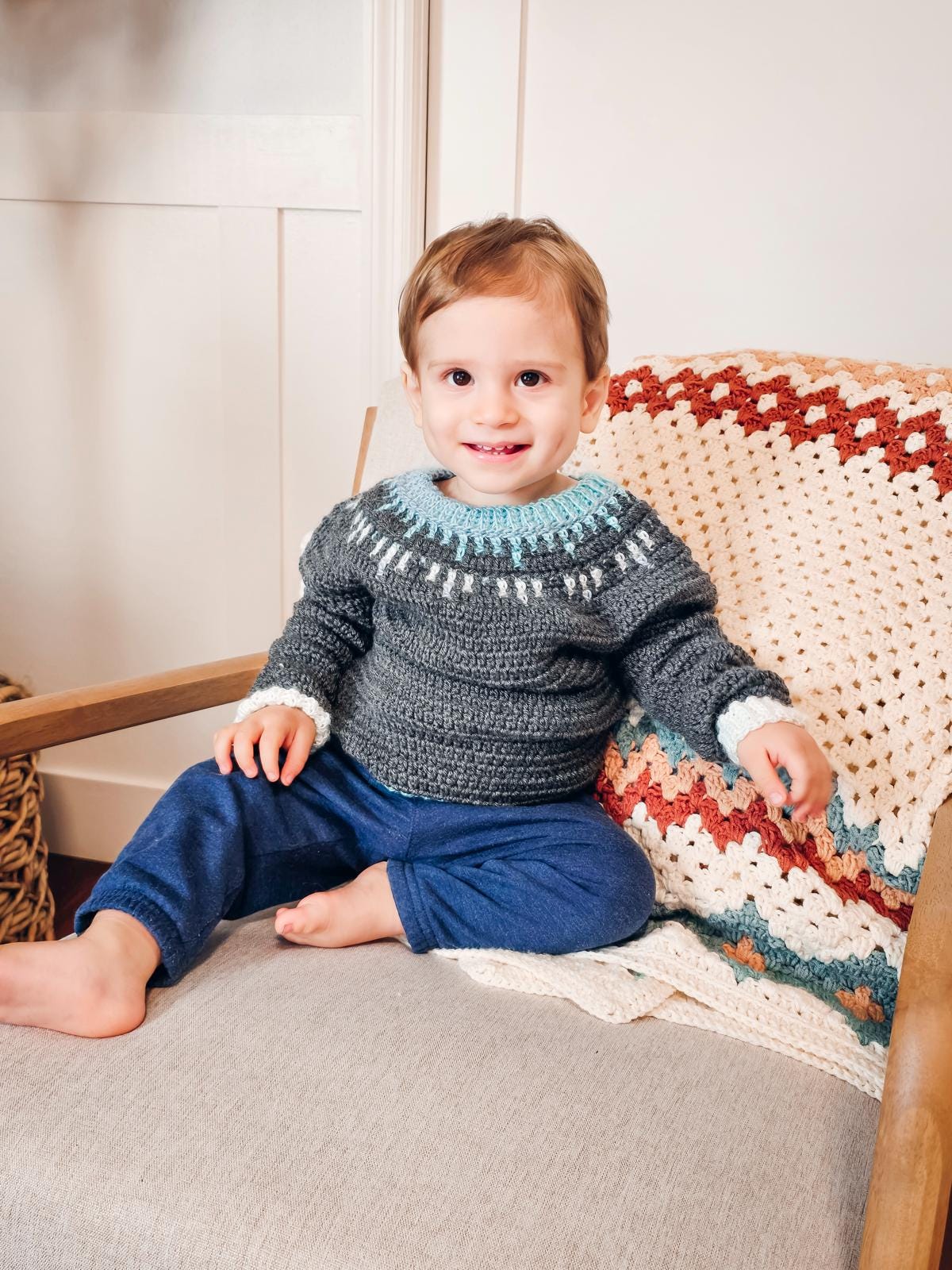 A child sits on a chair wearing a knitted sweater and blue pants, exuding the charm of vintage clothes. Nearby, a crocheted blanket acts as a delightful handmade gift, adding to the cozy ambiance.