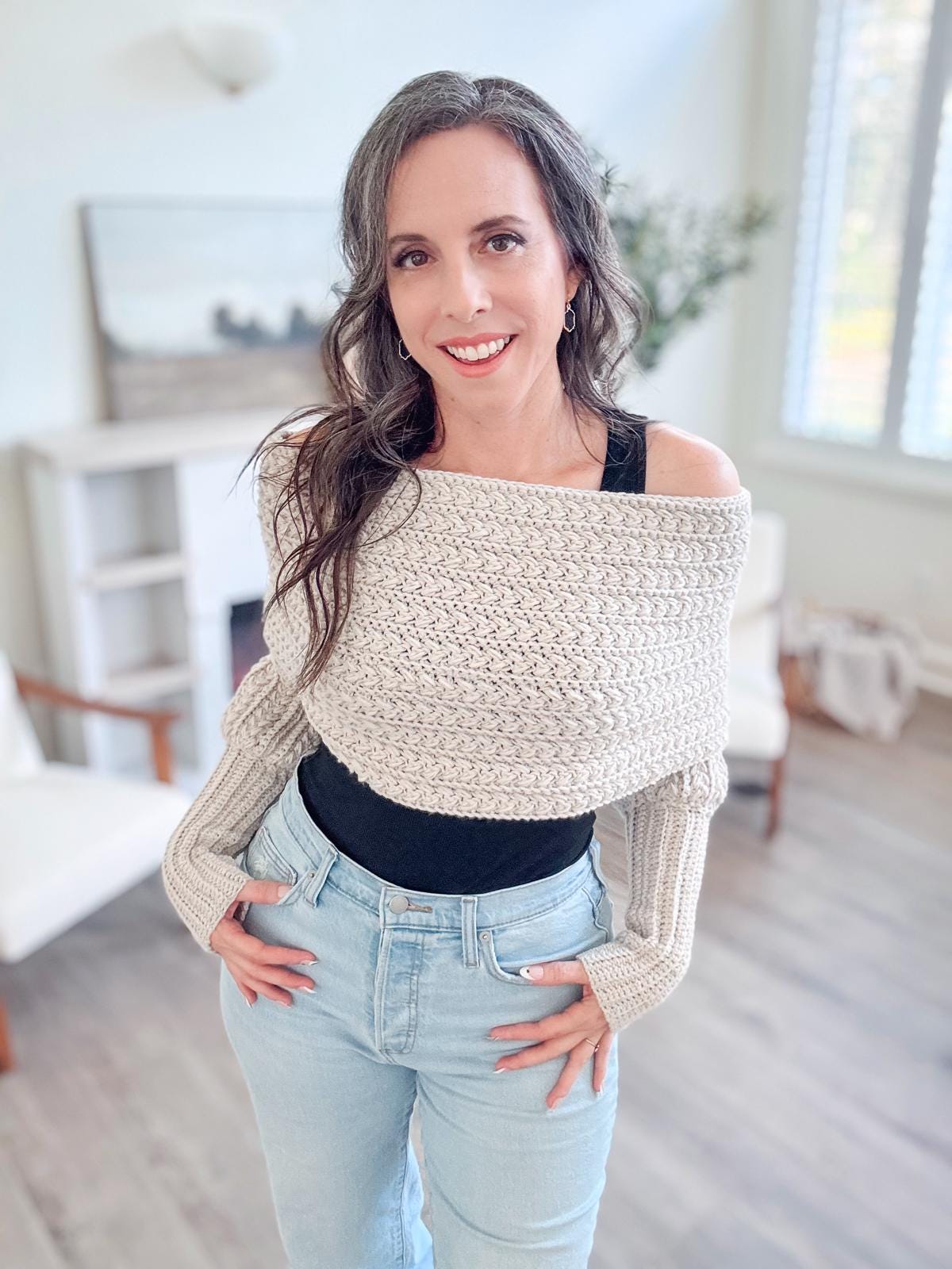 A woman with long wavy hair smiles, wearing a off-shoulder sweater adorned with unique jewelry and jeans, standing in a bright room with a window and minimal decor.