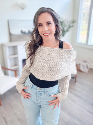 Cream crochet sweater scarf worn off the shoulders by a smiling woman in a black top and light jeans