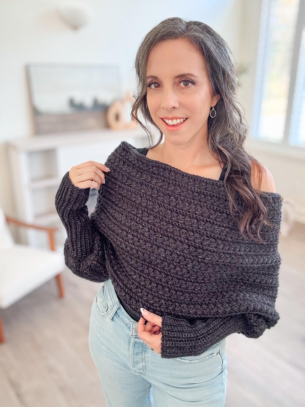 A woman stands indoors wearing a textured black  crochet sweater scarf and light jeans, smiling slightly while holding one side of the sweater. Her unique jewelry gleams subtly against the crochted fabric.