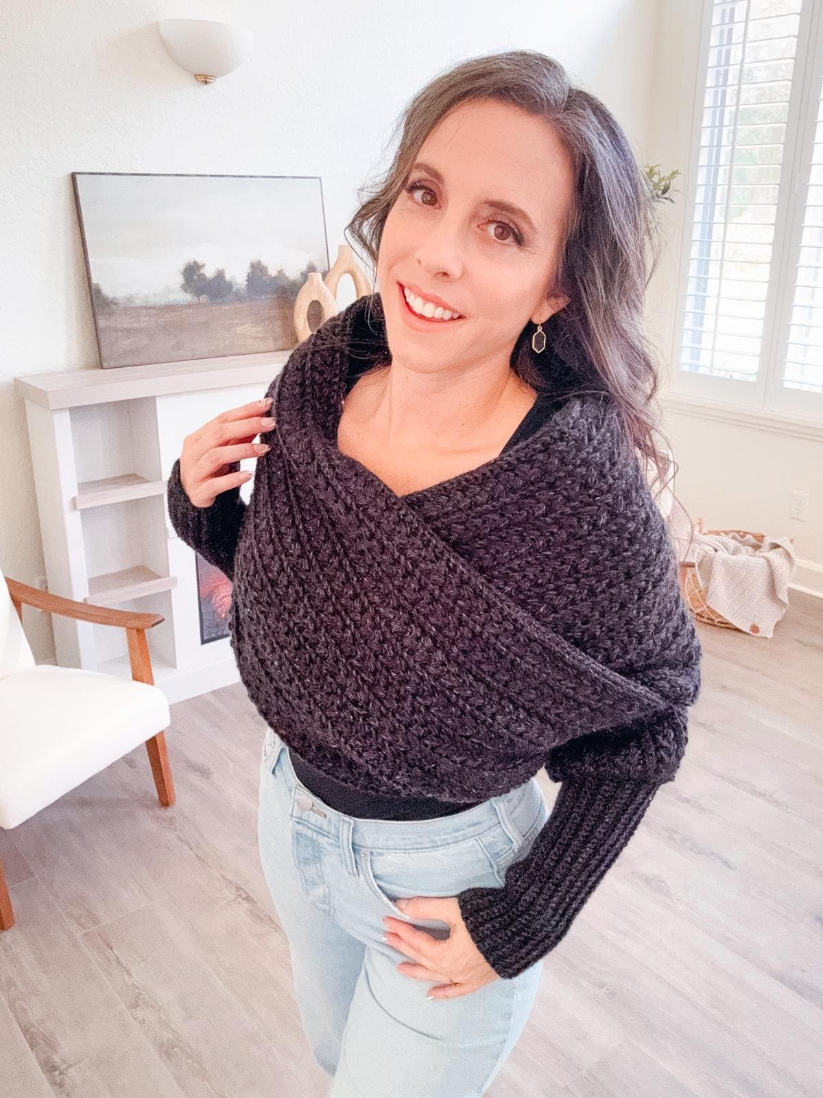 A woman stands indoors wearing a black crochet shawl and jeans, smiling at the camera. A chair, a small shelf with unique jewellery, and a framed picture are in the background.