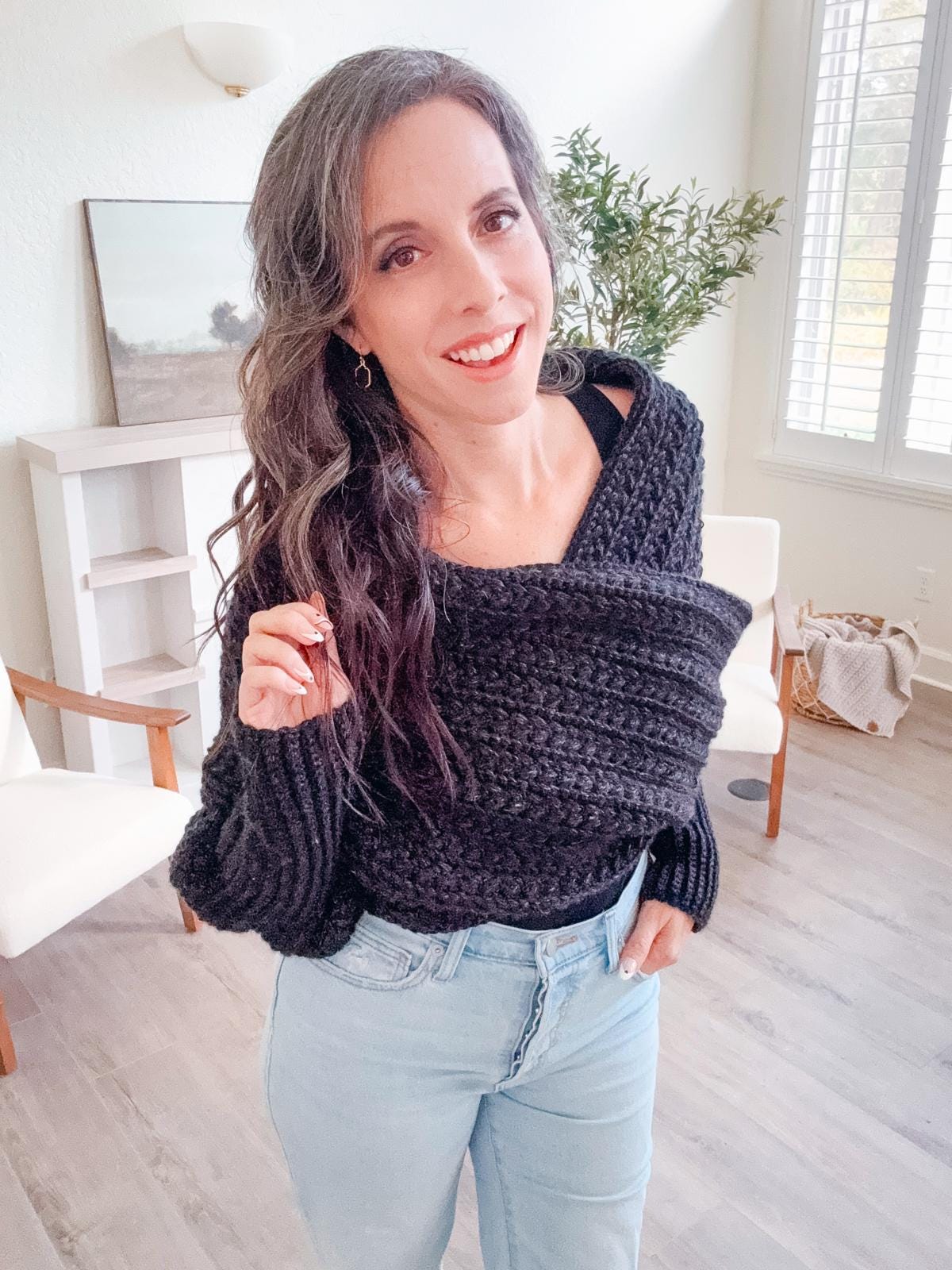 A woman stands indoors wearing a black crochet shawl and jeans, smiling at the camera. A chair, a small shelf with unique jewellery, and a framed picture are in the background.