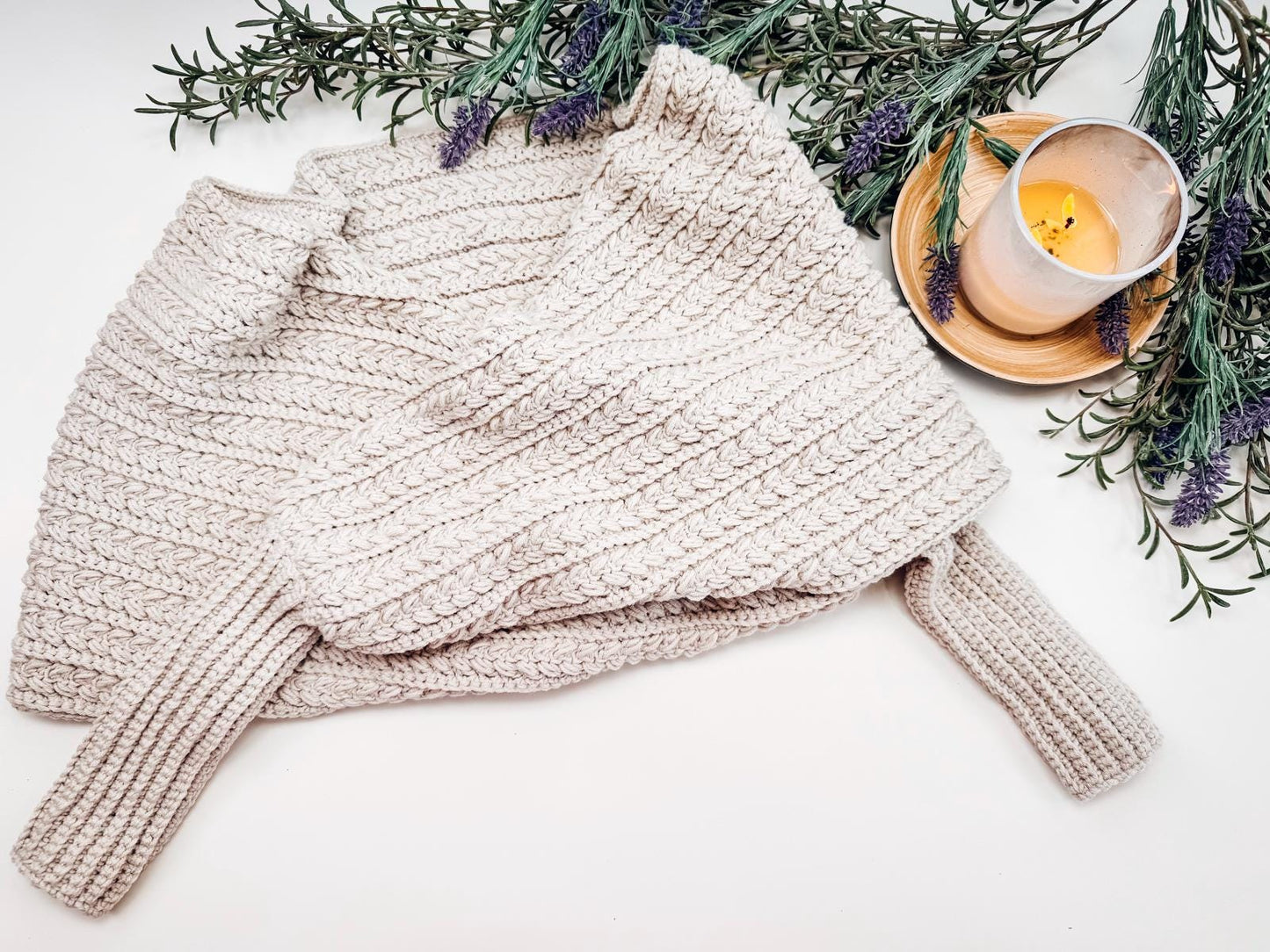 A beige crocheted sweater scarf is folded on a surface, surrounded by sprigs of lavender and a lit candle in a glass holder on a wooden tray.