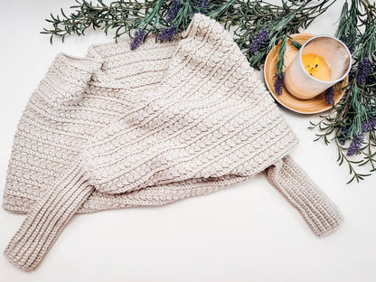 A beige crocheted sweater scarf is folded on a surface, surrounded by sprigs of lavender and a lit candle in a glass holder on a wooden tray.