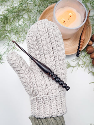 Light gray crochet mitten with braided texture worn on a hand, with a wooden crochet hook resting on top